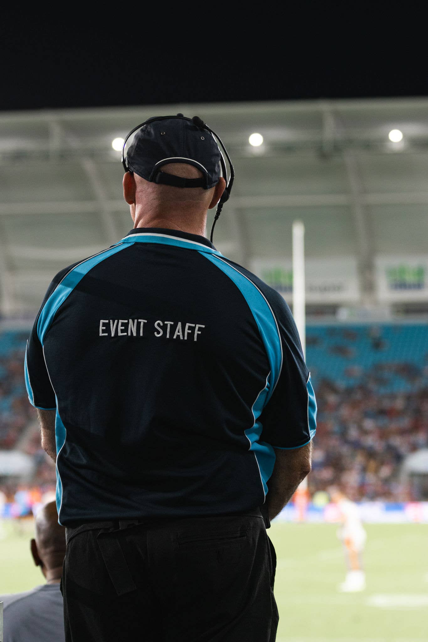 A man looks over a playing field wearing a headset and a shirt that reads "event staff"